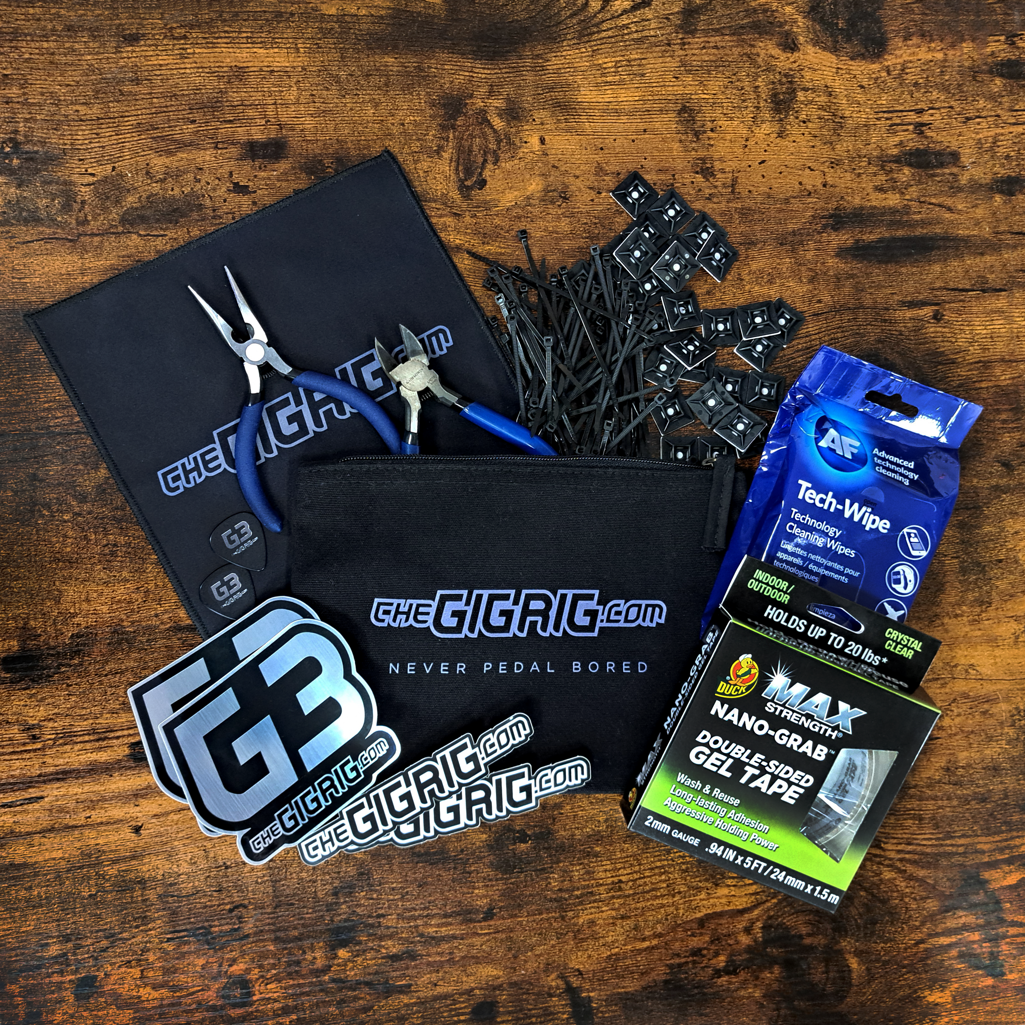 The GigRig Branded Organic pouch, Cable tes, cable tie mounts, G3 and GigRig Logo stickers, microfibre cleaning cloth, flush cutters, pliers, Technology cleaning Wipes, Nano Grab Double sided gel tape laid out on wooden table.
